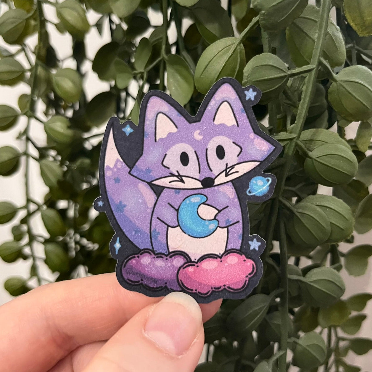 Sticker of Cosmic purple Maple the Fox sitting on some pink and purple clouds holding a little moon.