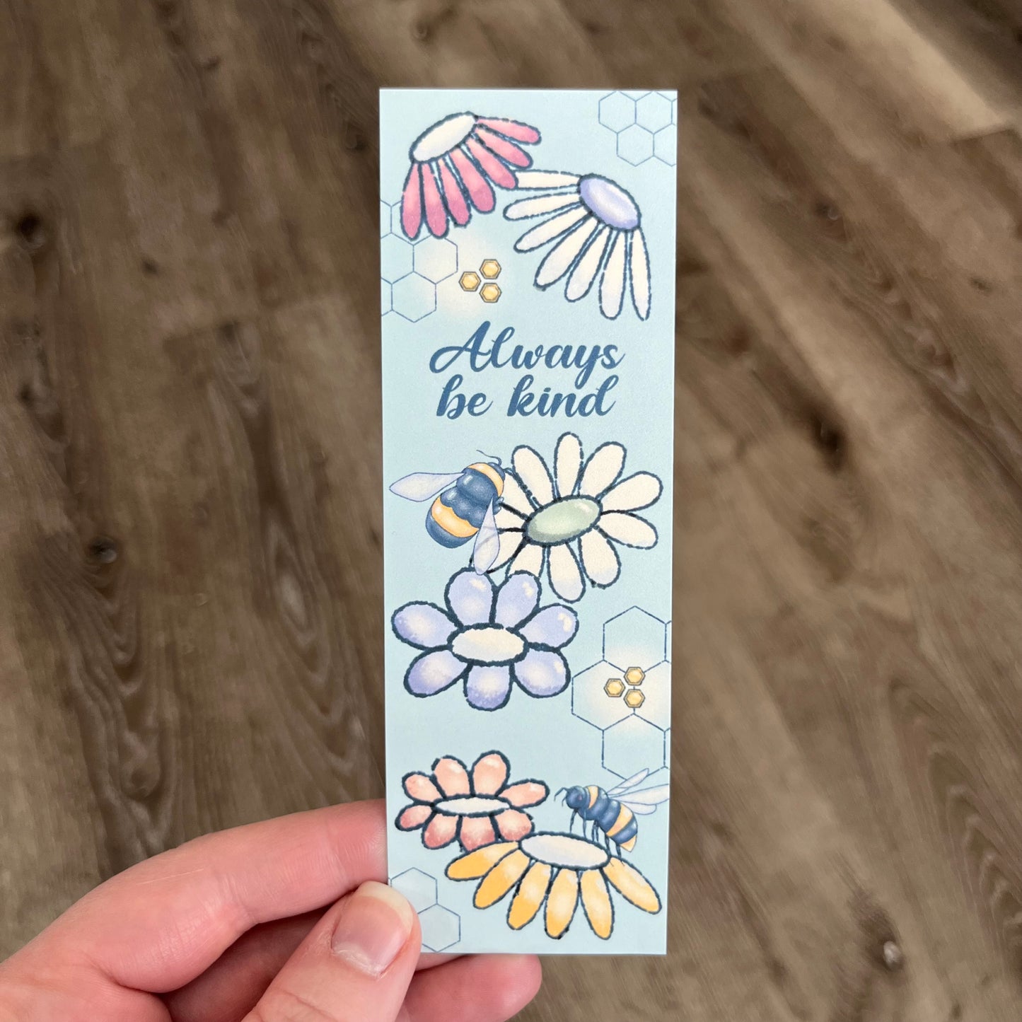 Bookmark with floral designs and 'Always be kind' text held against a wooden background