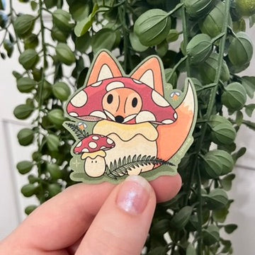 Die-cut compostable sticker of Maple the Fox dressed in a mushroom costume standing amongst ferns in the forest next to his mushroom pal. They are looking at fireflies.