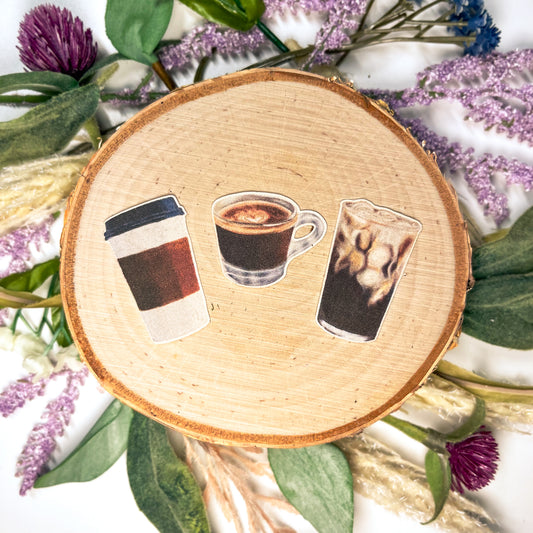3 Mini Coffee Stickers featuring hand drawn Espresso, Iced Coffee, and a To-Go Cup sitting on top of a wooden display surrounded by flowers